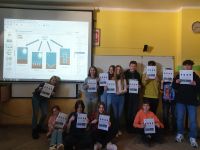 Grupa uczniów stoi przy ścianie z tablicą multimedialną, w ręce trzymają swoje prace