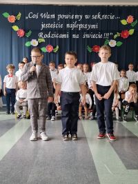 Trzech chłopców stoi na tle grupy, jeden trzyma mikrofon w ręce