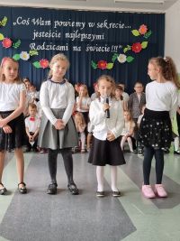 Z przodu stoją cztery dziewczynki, najmniejsza z nich mówi do mikrofonu