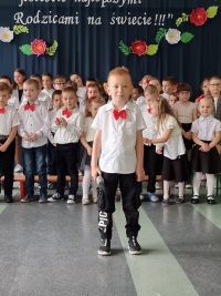 Chłopiec w biało-ciemnym stroju z czerwoną muchą stoi na tle grupy